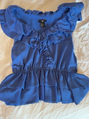 Blå volangblus från H&M - Supersöt blå blus från H&M med volanger längs ringningen och nederkanten. Blusen är ärmlös och har en luftig, flowy passform med resår i midjan. Perfekt för dig som gillar romantiska detaljer och en feminin look.