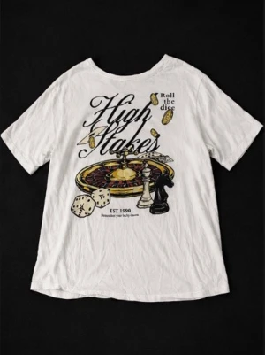 Vit t-shirt med casino-tryck - Vit t-shirt med coolt casino-inspirerat tryck på ryggen med texten 'High Stakes', tärningar, schackpjäs och roulettehjul. T-shirten har rund hals och korta ärmar. Perfekt för dig som gillar unika prints och streetstyle. Aldrig använd helt ny💥💥