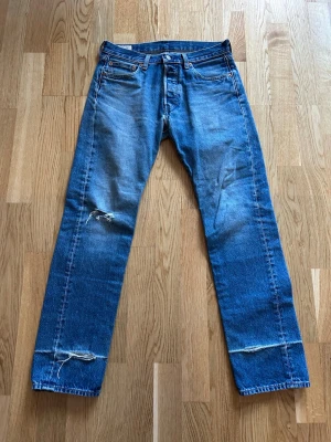 Blå raka 501 jeans från Levi's - Klassiska blå jeans från Levi's med rak passform och slitna detaljer vid knä och benslut. Jeansen har fem fickor, knappgylf och tydliga slitningar som ger en cool vintagekänsla. Strl W30 L32.