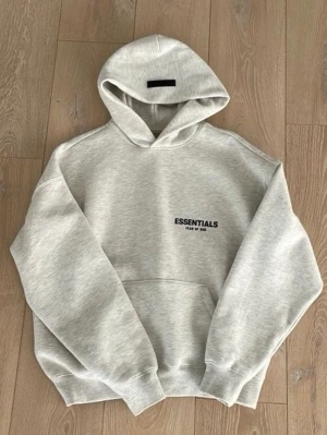 Essentials Fear of God hoodie grå -  Intresse koll på en Grå hoodie från Essentials Fear of God med svart logga på bröstet och stor text på ryggen. Klassisk passform med huva och känguruficka. 