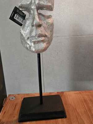 Dekorativ silverfärgad ansiktsmask - Unik dekoration i form av en silverfärgad ansiktsmask på svart metallfot. Masken har en konstnärlig, halvöppen design och står stadigt på en rektangulär bas. Perfekt som cool inredningsdetalj i vardagsrum eller sovrum.