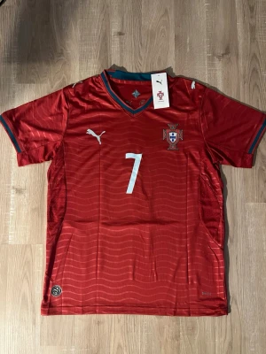 Portugal Ronaldo VM-2026 Fotbollströja - Röd Portugal fotbollströja med nummer 7 och Ronaldo på ryggen. Tröjan har korta ärmar, gröna och röda detaljer vid kragen och är tillverkad i ett lätt, ventilerande material. Perfekt för fotbollsträning eller att bara bära som supporter. HELT NY
