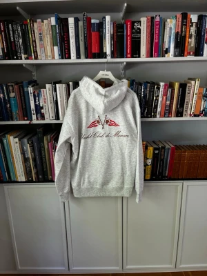 Julian Vand grå hoodie Yacht Club de Monaco - Snygg ljusgrå hoodie från Yacht Club de Monaco med tryck av två röda flaggor och text på ryggen. Hoodien har huva och ribbade muddar vid ärmslut och nederkant. Perfekt för en chill och sportig look.