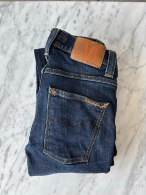 Nudie Jeans - Snygga mörkblå jeans från Nudie Jeans i modellen grimtim med klassisk femficksmodell och kontrastsömmar. Jeansen har en rak passform och är tillverkade i slitstark denim med läderpatch bak i midjan. Perfekta för dig som gillar stilrena och tidlösa jeans.