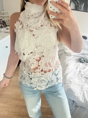 Vit spetsig halterneck topp - Säljer en superfin vit halterneck topp i spets med hög krage och öppen rygg. Toppen knyts i nacken med breda band och har ett genombrutet blommigt mönster. Perfekt för dig som vill sticka ut med en unik och elegant look. storlek xs-m
