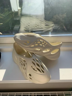 Beige Yeezy Foam Runner tofflor - Säljer ett par beige Yeezy Foam Runner tofflor med futuristisk design och många ventilationshål. Tillverkade i lätt och bekväm EVA-skum som formar sig efter foten. Perfekta för chill dagar och streetwear-stil.