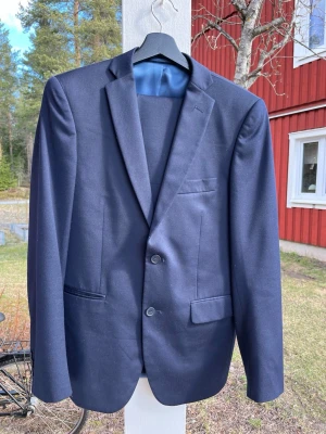 Mörkblå slim fit kostym 46 - Studentkostym! Snygg mörkblå kostym från Dressmann i slim fit-modell. Kavajen har två knappar framtill, klassiska fickor och en bröstficka. Perfekt för dig som vill ha en stilren och modern look. Fodrad insida i blått tyg. Mörkblå kostym från Dressman i storlek 46 Slimfit  midjemått: 85cm Beninnerlängd: ca 82 cm  Rygglängd från Nacke och ner: 72cm Bredd över rygg, ärmhål till ärmhål ca 54cm  Bra skick  Rök- och djur-fritt hem