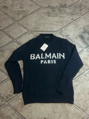 Mörkblå stickad tröja Balmain Paris - Snygg mörkblå stickad tröja från Balmain Paris med vit logotyp framtill. Tröjan har rund halsringning och ribbstickade muddar vid ärmslut och nederkant. Perfekt för dig som gillar exklusiva och trendiga plagg.