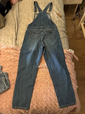 Blå denim hängselbyxor - Säljer ett par klassiska blå denim hängselbyxor med raka ben och flera fickor både fram och bak. Bäst av allt DE ÄR HELT NYA! Byxorna har justerbara axelband med metallspännen och är tillverkade i slitstarkt bomullsjeansmaterial. Perfekta för en avslappnad och trendig look.
