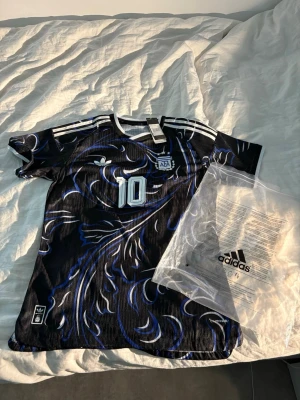 Svart Adidas fotbollströja Argentina vem  - Snygg svart fotbollströja från Adidas med blått och vitt mönster, nummer 10 och Argentina-emblem på bröstet. Tröjan har klassiska Adidas-ränder på axlarna och är tillverkad i lätt funktionsmaterial. Perfekt för dig som älskar fotboll och vill sticka ut på planen. Passar M 