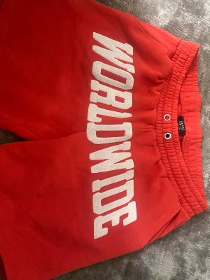 WORLDWIDE - Röda shorts från bohooman med stort vitt tryck 'WORLDWIDE' framtill. Shorts med resår i midjan och dragsko för skön passform. Perfekta för sommardagar eller avslappnade tillfällen.