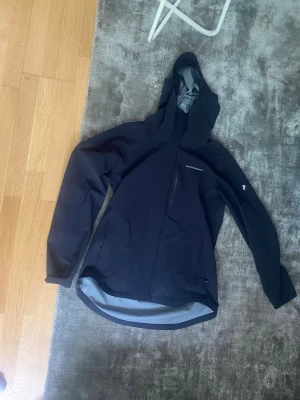 Svart regnjacka från Peak Performance - Snygg svart regnjacka från Peak Performance med huva och dragkedja framtill. Jackan har vattentäta fickor och logga på bröstet samt ärmen. Perfekt för regniga dagar och outdoor-äventyr.