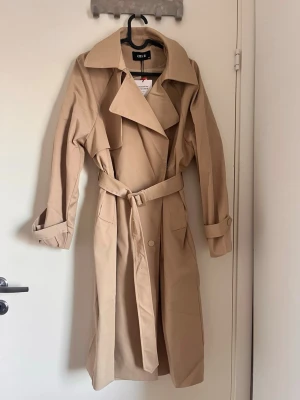 Beige trenchcoat från Cider  - Den är oanvänd. Jag köpte den och tyckte den va lite stor på mig (jag har storlek XS). Skulle säga att den passar mer storlek S. Färgen på kappan när jag tog bilder såg lite mörkare ut på grund av ljuset men det är den färger som är från cider den som stämmer. Nypris är 1000kr. Har du några frågor skriv till mig