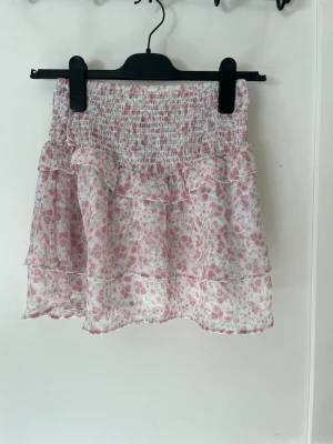 Blommig volangkjol i rosa och vitt - Söt kortkjol med blommigt mönster i rosa och vitt. Kjolen har smock i midjan och dubbla volanglager.