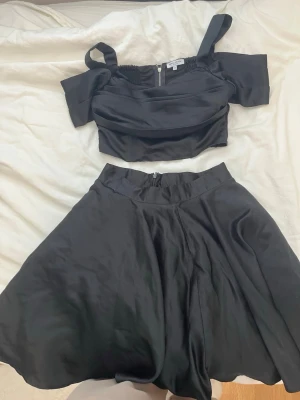 Svart satinsätt med topp och kjol - Snyggt svart satinsätt från Bubbleroom med en crop top och matchande vid kjol. Toppen har breda axelband och smock i ryggen för skön passform. Kjolen är högmidjad och har dragkedja bak. Perfekt för en trendig look.