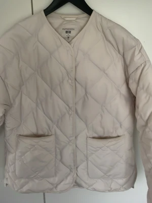 Ljusbeige pufferjacka från Uniqlo - Stilren ljusbeige pufferjacka från Uniqlo med quiltat mönster och två stora framfickor. Jackan har tryckknappar och en enkel, kraglös design. Perfekt för kalla dagar och lätt att matcha med olika outfits.