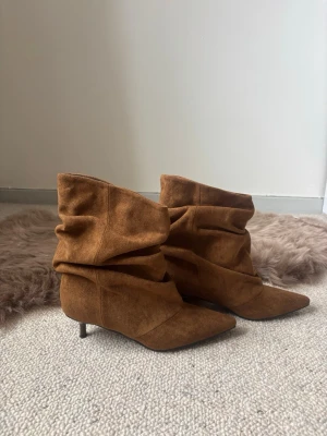 Bruna slouchy stövletter i mocka - Säljer ett par bruna stövletter med slouchy design och spetsig tå. Skorna har smal klack och är gjorda i mjuk mocka som ger en trendig och avslappnad look. Perfekta för dig som vill ha något snyggt och bekvämt till hösten.