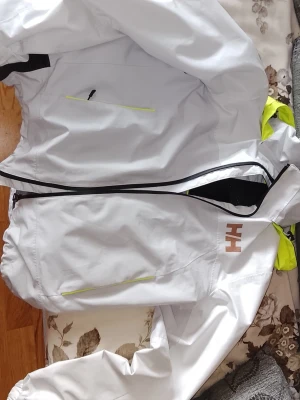 Vit vindjacka från Helly Hansen - Snygg vit vindjacka från Helly Hansen med svarta och neongul luva.. Jackan har används ett litet tag men har ej permanenta fläckar, repor elle så