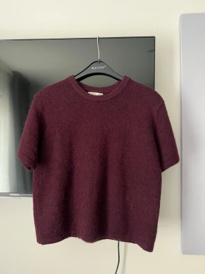 Vinröd stickad tshirt Gina Tricot - Mysig vinröd stickad tröja från Gina Tricot med korta ärmar och rund halsringning. Tröjan har en enkel, boxig passform och är gjord i ett mjukt, luddigt material som känns skönt mot huden. Perfekt för lager på lager och en avslappnad look.