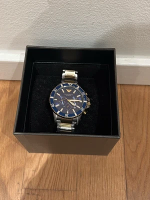 Emporio Armani blå-silver herrklocka - Stilren herrklocka från Emporio Armani med blå urtavla, silverfärgad länk och gulddetaljer. Klockan har tydliga index, roterande bezel och klassisk logga vid tolvslaget. Perfekt accessoar för dig som vill ha en elegant och modern look.