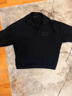 Svart Essentials hoodie med krage - Svart hoodie från Fear of God Essentials med klassisk krage och diskret logga på bröstet. Oversized passform och ribbade muddar nedtill. Perfekt för en clean och stilren streetwear-look.
