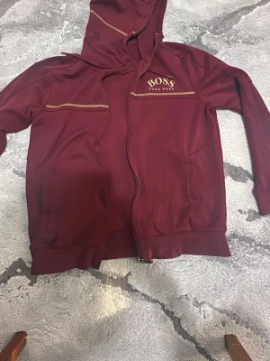 Vinröd hoodie från Hugo Boss - Snygg vinröd hoodie från Hugo Boss med dragkedja och huva. Har guldfärgade detaljer och logga på bröstet samt två fickor framtill. Perfekt för en avslappnad och trendig stil.