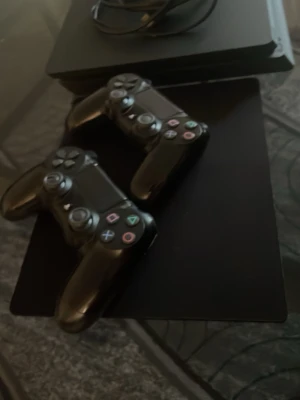 PlayStation 4 Slim med två handkontroller - Säljer en svart PlayStation 4 Slim med två trådlösa DualShock 4-kontroller och tillhörande laddningskabel. Konsolen har en stilren, matt finish och är perfekt för gamingkvällar med vänner. Allt är i klassisk svart plast och har den ikoniska PlayStation-designen.