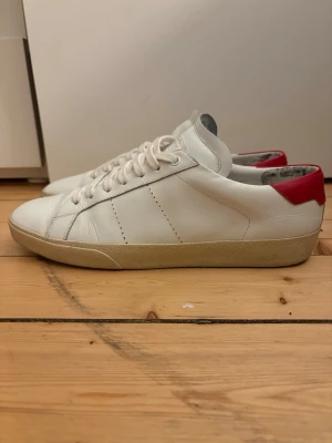 Saint laurent skor - Snygga vita sneakers från Saint Laurent i läder med röd detalj på hälen och beige sula. Klassisk låg modell med vita skosnören och diskreta sömmar. Perfekta för dig som gillar stilrena och trendiga skor. Box ingår