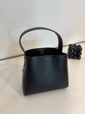 Svart bucket bag i läderlook - Svart handväska i bucket-modell med bredt handtag och minimalistisk design. Väskan har silverfärgade detaljer på sidorna och ett innerfack med dragkedja. Perfekt för dig som gillar enkel och modern stil.🤍