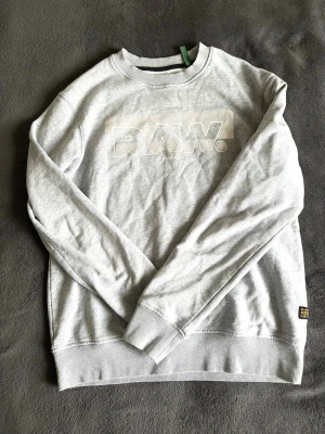 Grå sweatshirt från G-Star RAW - Snygg ljusgrå sweatshirt från G-Star RAW med broderad RAW-logga framtill och en svart/gul patch på ärmen. Tröjan har rund hals, ribbade muddar och är tillverkad i mjukt bomullsmaterial. Perfekt för en avslappnad streetstyle-look. Har dock synliga skador ( se bilder)