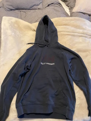 Mörkblå hoodie från Helly Hansen - Snygg mörkblå hoodie från Helly Hansen med klassisk logga på bröstet. Hoodien har stor huva med dragsko och en rymlig magficka. Perfekt för en avslappnad och sportig stil. Tillverkad i mjukt bomullsmaterial för extra komfort.