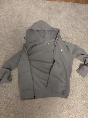 Grå hoodie från Polo Ralph Lauren - Snygg grå hoodie med dragkedja från Polo Ralph Lauren. Klassisk design med huva och dragsko, två fickor framtill och den ikoniska vita loggan broderad på bröstet. Perfekt för en chill och stilren look.