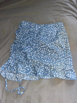 Blå blommig kortkjol med snörning - Söt blå kortkjol med vitt blommönster och volangkant. Kjolen har snörning på sidan som ger en snygg rynkad effekt. 