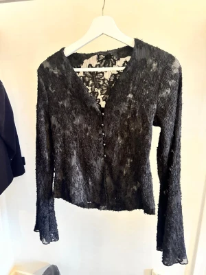 Svart transparent blus med spetsdetaljer - Snygg svart transparent blus med lång ärm och v-ringning. Blusen har dekorativa knappar framtill och spetsdetaljer vid halsen. Materialet är lätt och stretchigt. Använd en gång. Den har även lite utsvängda armar riktigt snyggt.