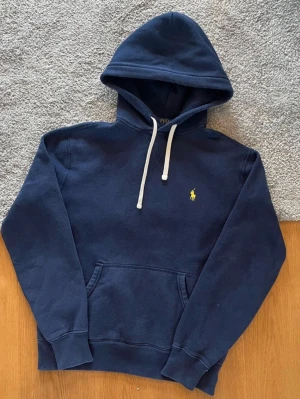 Mörkblå hoodie från Polo Ralph Lauren - Mörkblå hoodie från Polo Ralph Lauren med klassisk gul logga på bröstet. Hoodien har vit dragsko i huvan och en stor magficka framtill. Tillverkad i mjukt bomullsmaterial och har ribbade muddar vid ärmslut och nederkant. Perfekt för en avslappnad och stilren look. Passar folk runt 165 och neråt 