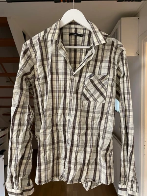 Rutig skjorta från Emilio, brun/vit - Säljer en långärmad rutig skjorta från Emilio i bruna, beige och vita toner. Skjortan har klassisk krage, bröstficka och knäppning framtill. Perfekt för dig som gillar avslappnad men ändå stilren look.