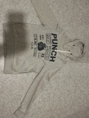 Ljusgrå hoodie med tryck från Punch - Ljusgrå hoodie från Punch med stort mörkblått tryck på bröstet och text om boxing club. Klassisk huva med vita dragsnören och magficka framtill. Tillverkad i mjukt bomullsmaterial och har ribbade muddar vid ärmslut och nederkant. Perfekt för en avslappnad streetstyle.