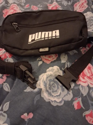 Svart magväska från Puma - Snygg svart magväska från Puma med justerbart band och plastspänne. Väskan har ett stort huvudfack och en mindre ficka framtill med dragkedja. Tillverkad i slitstarkt tyg med Puma-logga i vitt på framsidan. Perfekt för att bära det viktigaste på ett smidigt sätt.