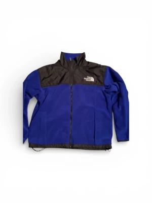 Blå fleecejacka från The North Face - Blå fleecejacka från The North Face med svarta detaljer över axlar och bröst. Jackan har hel dragkedja, hög krage och två sidofickor. Materialet är mjuk fleece och partierna upptill är i slitstark nylon. Perfekt för kyliga dagar. Kan användas som skid jacka.