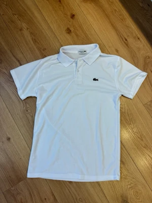 Lacoste pikétröja - Klassisk vit pikétröja från Lacoste med korta ärmar och krage. Tröjan har två knappar framtill och den ikoniska krokodilloggan broderad på bröstet. Regular fit och tillverkad i bomull för en skön känsla.