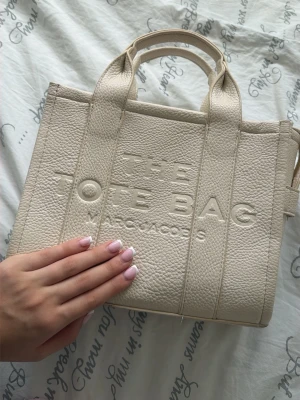 Beige Marc Jacobs tote bag - Säljer en beige shoppingväska från Marc Jacobs i präglat läder med texten 'THE TOTE BAG' på framsidan. Väskan har dubbla handtag och en stilren, boxig form. Perfekt för dig som vill ha en trendig och rymlig tote i vardagen.