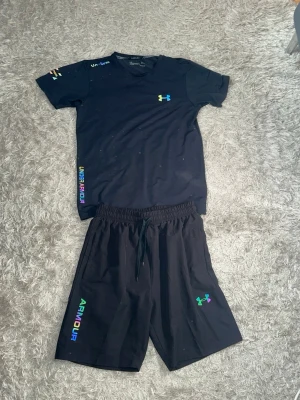 Svart Under Armour set med regnbågsdetaljer (reflex) - Säljer ett svart set från Under Armour med t-shirt och shorts. Båda plaggen har logga och text i regnbågsfärger (reflex). T-shirten har rund hals och kort ärm, shortsen har dragsko i midjan. Perfekt för en sportig och avslappnad stil.