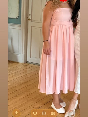 Ljusrosa långklänning med knytband - Söt ljusrosa långklänning med smockad överdel och axelband som knyts i rosetter. Klänningen har en utsvängd kjol och fyrkantig ringning. Perfekt för sommarens alla tillfällen.