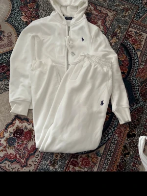 Polo Ralph Lauren hoodie med byxorna - Snygg vit hoodie från Polo Ralph Lauren med dragkedja framtill och klassisk huva med dragsko. Tröjan har två fickor på magen och den ikoniska blå loggan broderad på bröstet. Tillverkad i mjukt material som känns skönt mot huden. Perfekt för en avslappnad och stilren look. Funkar för män och kvinnor, storleken är M men funkar lika bra för S, aldrig använt. 