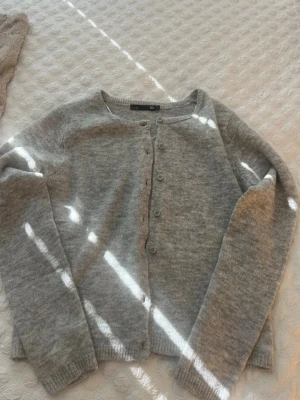 Ljusgrå kofta från 157 - Mysig ljusgrå kofta från 157 med rund halsringning och knäppning framtill. Tillverkad i mjukt stickat material och har långa ärmar samt ribbade muddar vid ärmslut och nederkant. Perfekt för lager på lager och enkel att matcha.