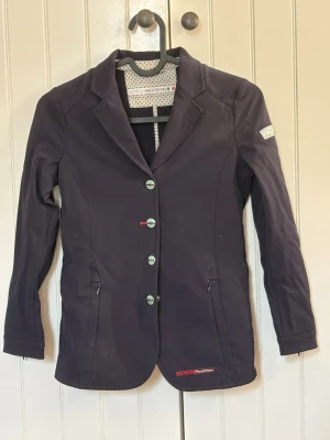 Marinblå kavaj från Animo - Snygg marinblå kavaj från Animo med figurnära passform, klassisk krage och fyra knappar framtill. Två dragkedjefickor och diskret logga på ärmen samt vid nederkanten. Fodrad med prickigt tyg för extra stil. Storlek 140 barn