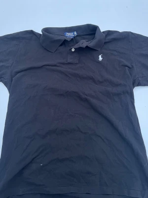 Polo Ralph Lauren Piké  - SITTER SOM M,    Klassisk svart pikétröja från Polo Ralph Lauren med korta ärmar och vit broderad logga på bröstet. Tröjan har krage och två knappar vid halsen. Tillverkad i mjuk bomull som känns skön mot huden. Perfekt för en avslappnad men ändå stilren look.