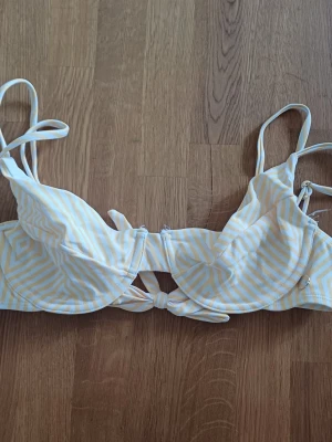 Randig bikiniöverdel med knyt framtill - Snygg bikiniöverdel med vita och ljusgula ränder. Modellen har bygel, smala axelband och en dekorativ knytning framtill. Perfekt för soliga dagar på stranden. Materialet känns mjukt och bekvämt mot huden.