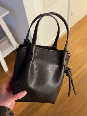 Svart bucket bag i skinn - Snygg svart bucket bag i slätt skinn med två handtag och dekorativ knutdetalj på sidan. Väskan har en stilren och minimalistisk design som passar till många olika looks. Perfekt storlek för det viktigaste och enkel att bära med sig.