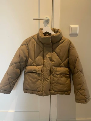 Beige quiltad pufferjacka - Säljer en beige pufferjacka med quiltat mönster. Jackan har två stora fickor framtill med lock och tryckknappar samt dragkedja och tryckknappar för stängning. Perfekt för kalla vinterdagar och riktigt skön att bära.